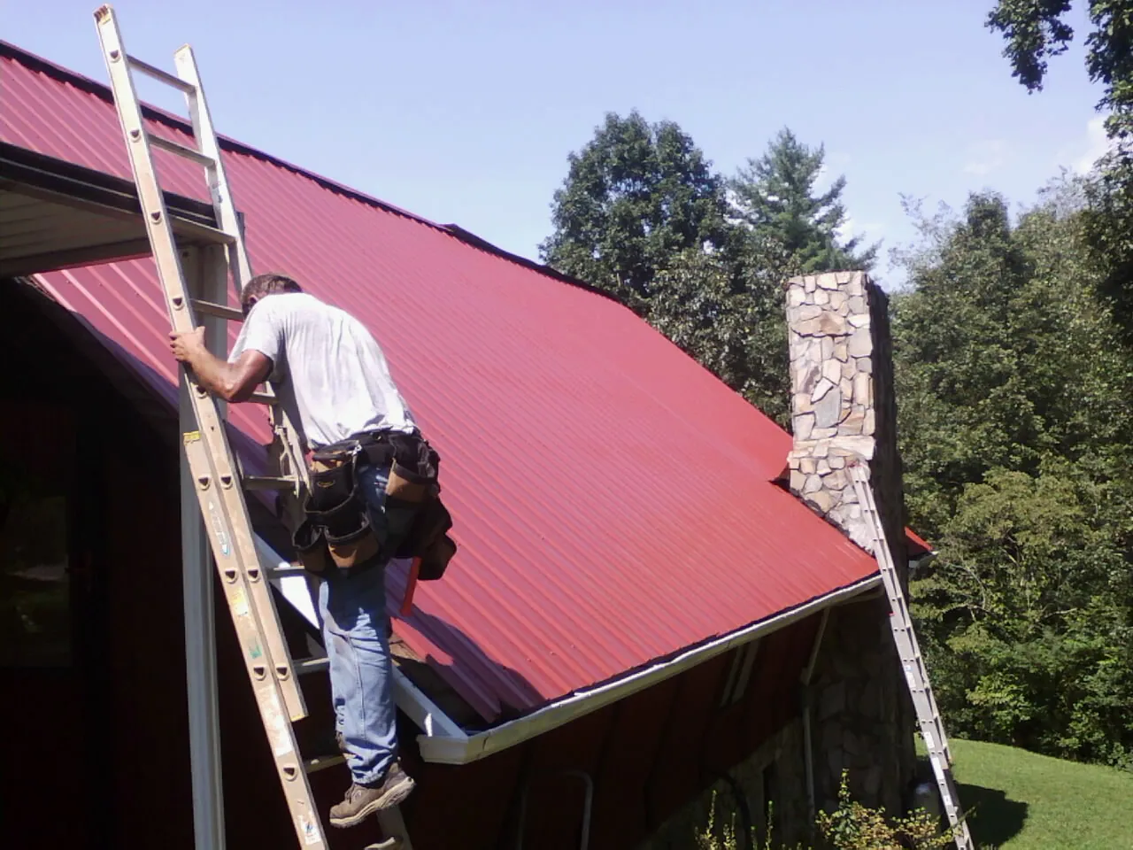 Roofer on a ladder beside a red metal roof and stone chimney — Soffit & Fascia Repair by Roofing Brothers in Elmhurst